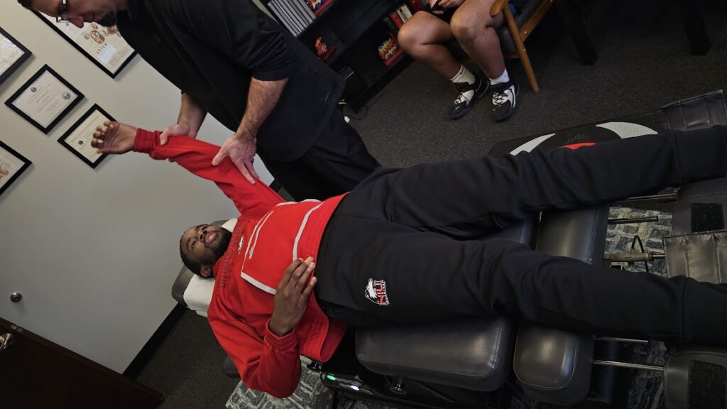 Man in red shirt receiving chiropractic adjustment on therapy bench at clinic gym.