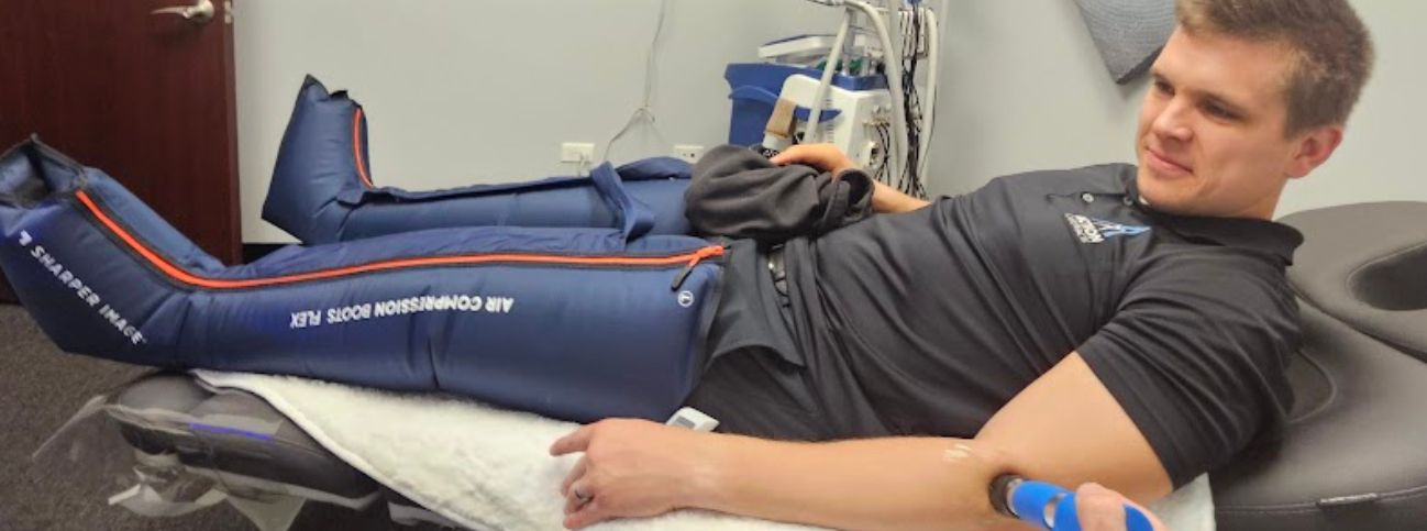 Man lying on a treatment table wearing blue air‑compression boots during leg therapy in a clinical setting.